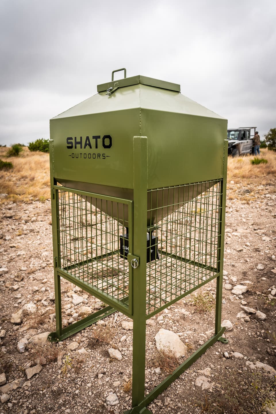 Three-quarter view of the whole feeder on rocky ground: hopper, lid handle, caged feed area with a pinned door, and legs on ground rails.