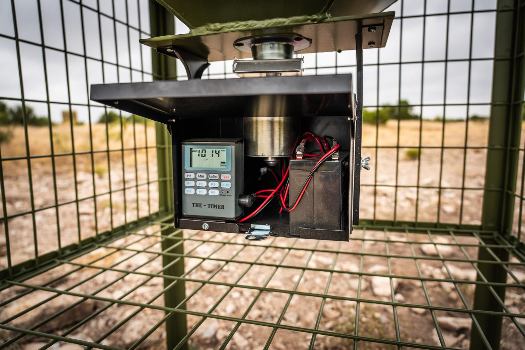 The open control box inside the cage, showing the digital timer, the spinner motor housing and a 12V battery with red and black leads.
