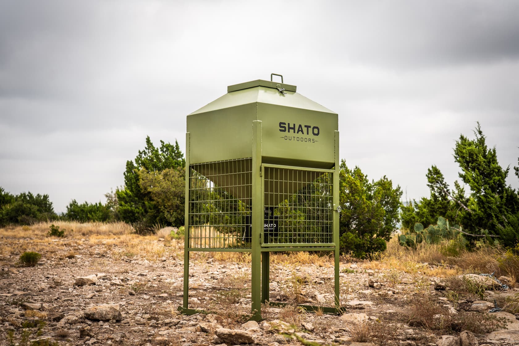 The whole Shato Outdoors feeder on rocky ground: olive powder-coated hopper with a lid handle, welded-wire cage with a door, and legs on ground rails.