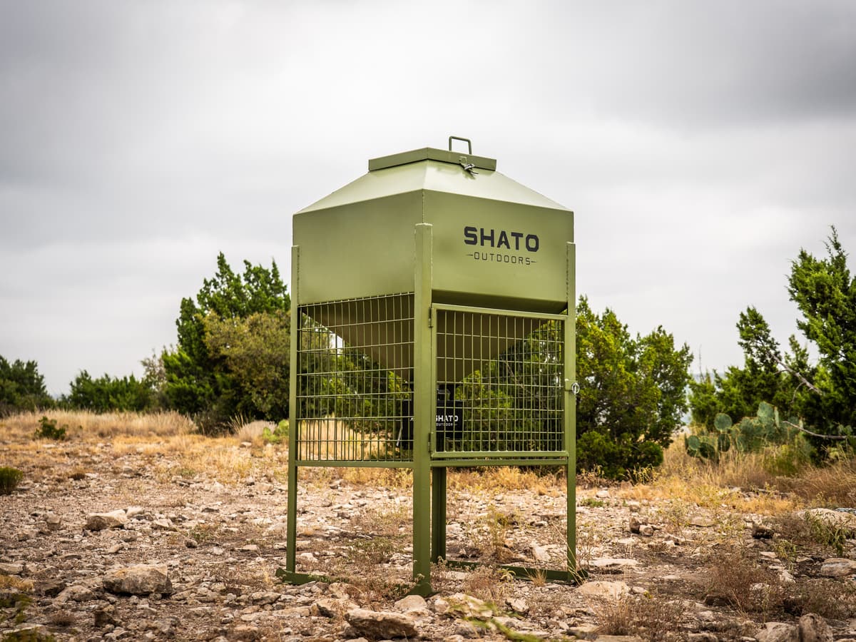 The whole Shato Outdoors feeder on rocky ground: olive powder-coated hopper with a lid handle, welded-wire cage with a door, and legs on ground rails.