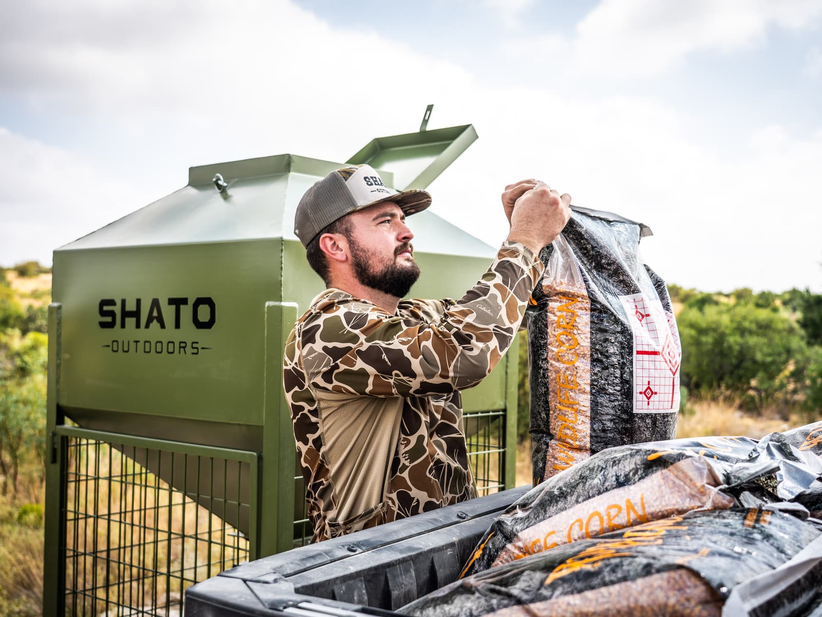 A man in a camo shirt and Shato cap lifts a bag of wildlife corn from a utility vehicle beside the feeder.