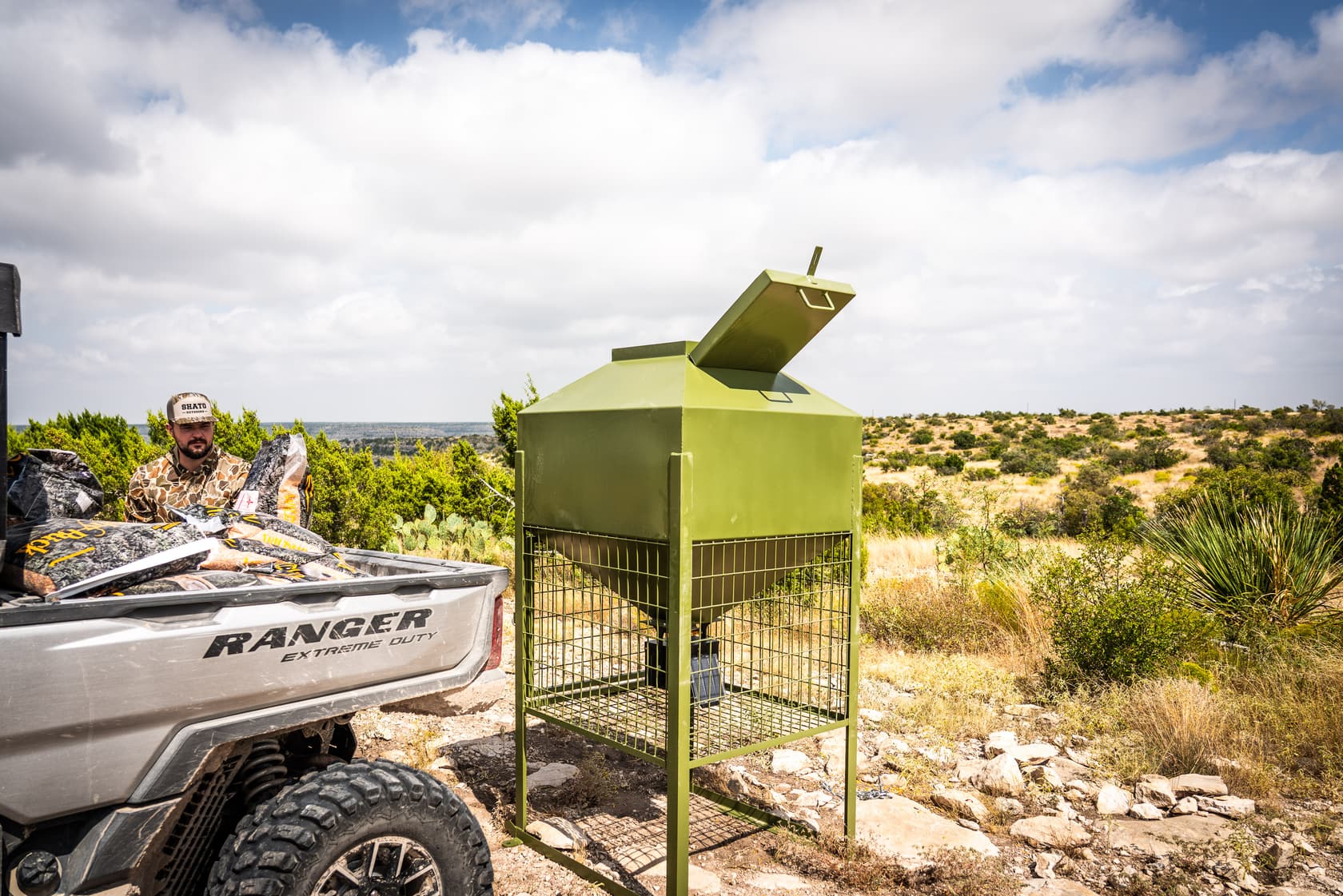 A utility vehicle loaded with feed bags parked beside the feeder, lid open, on a rise above West Texas brush country.